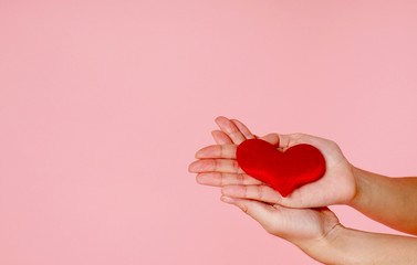 two hands holding a small red heart in palms on the pink background