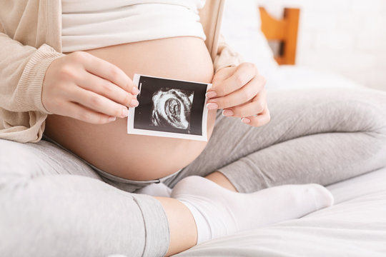 Pregnant woman holding ultrasound picture near belly, closeup