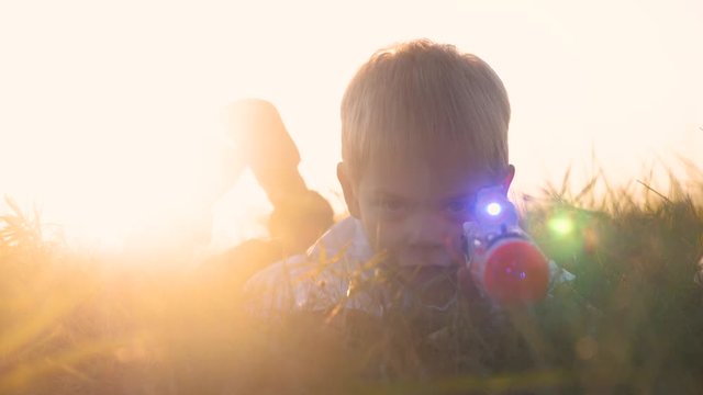 boy shoots from toy weapons gun. little boy plays war lies on the ground shoots an automatic blaster weapon of the future. slow motion sunset silhouette children lifestyle and weapons war concept