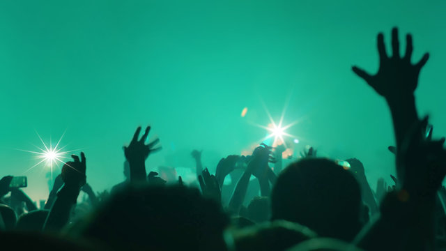 Photo of a concert hall with people silhouettes clapping in front of a big stage lit by spotlights. Shot is taken from concert crowd point of view, lens flare is visible