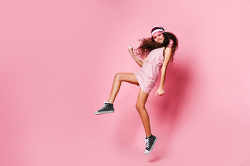 Teenager girl in a cap and pink striped dress is jumping joyfully shows a winner luck sign on a pink with copy space