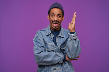 Excited young bearded brunette guy with dark skin raising hand while looking emotionally at camera with wide mouth opened and frowning his face, isolated over purple background