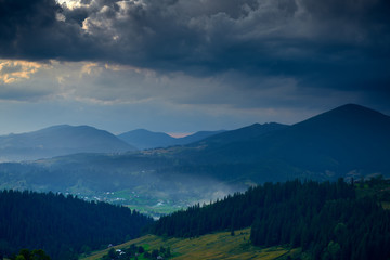 Sunset in carpathian mountains - beautiful summer landscape, spruces on hills, village, homes, dark cloudy sky and bright sun light, meadow and wildflowers