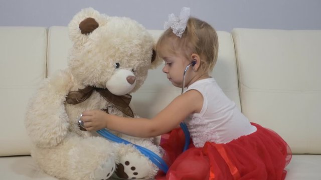 Little girl plays a doctor with a stethoscope and a teddy bear. girl sitting on the couch. The concept of children's fantasies and games. Training as a doctor