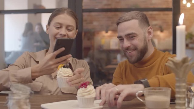 Close up of young Caucasian man and woman sitting in cafe and making photos of delicious cupcakes using phones camera
