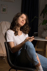 Obraz premium Curly hair girl in casual clothes sitting on chair at home with her phone in hand