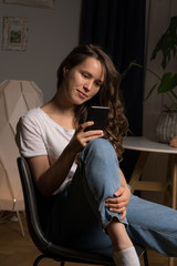 Curly hair girl in casual clothes sitting on chair at home with her phone in hand