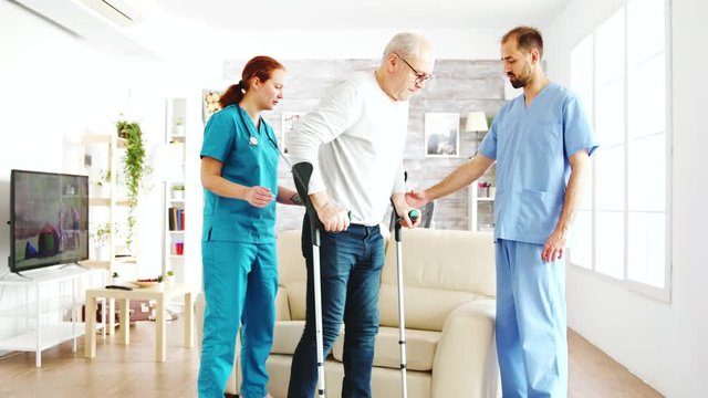 Two nurses hekping an old disabled man with crutches to walk in his room