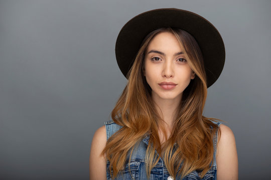 Beautiful hipster girl in stylish hat and denim vest posing at grey copy space wall for your content.