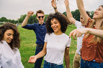 Group of friends at the park dancing under a rain of confetti - Millennials have fun in a public garden in summer at sunset - 319258512