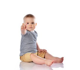 Infant baby boy in t-shirt and pants is sitting on the floor pointing at something up, pushing on white with copy space