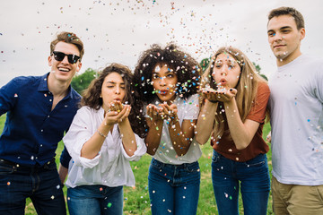 Group of four friends having fun at the park blowing confetti - Millennials playing together at...