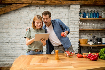 Lovely cheerful young couple cooking dinner together, looking recipe at digital tablet and having fun at rustic kitchen