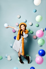 Fototapeta premium Asian child in oversized shirt dress, brown beret and scarf, boots. She sending air kiss, posing with balloons on blue background.