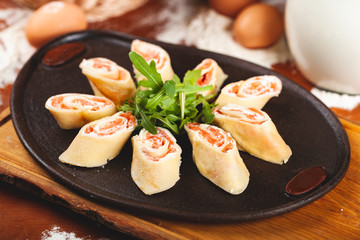 Rolled pancakes (crepes, blini) with smoked salmon and cream cheese served with arugula on black plate.