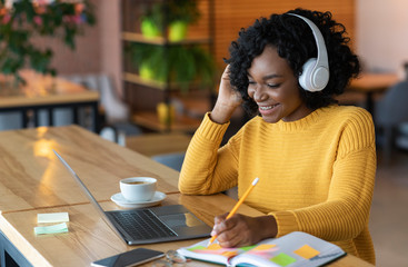 Smiling african girl in headphones looking at laptop, cafe interior