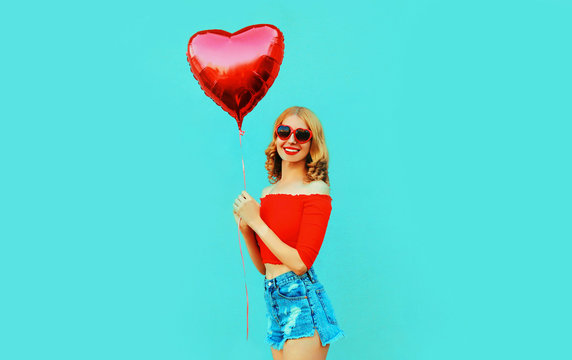 Portrait cute happy smiling woman in shorts holding red heart shaped air balloon on colorful blue background