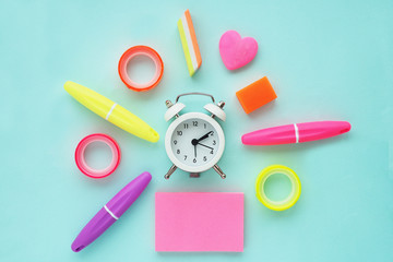 White alarm clock in the center and around it like sunbeams stationery: markers, erasers, colorful tape, pink stickers. Flat layout, top view on a blue background.