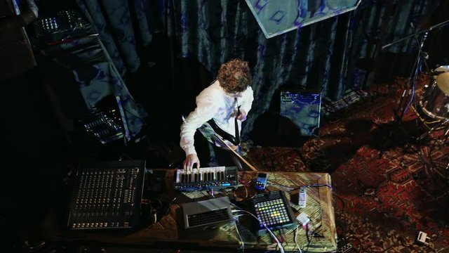 Musician in white shirt plays bass guitar and midi controller in the club with neon lights form above.