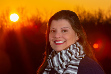 Lady outdoor closeup portrait  during a beautiful cold colorful winter sunset. She is smiling and looking at the camera. She is wearing season clothes.