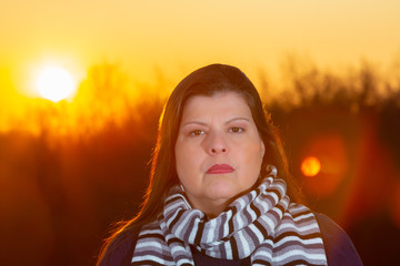 Lady outdoor closeup portrait  during a beautiful cold colorful winter sunset. She is serious, looking at the camera and wearing season clothes.