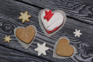 Marshmallows sandwiches on a decorative napkin. Nearby are gingerbread cookies in the shape of a heart and decorative snowflake cookies. On pine brushed boards painted in black and white.