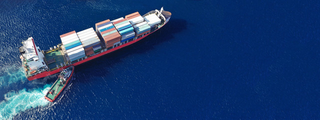 Aerial drone ultra wide top down photo of container ship cruising in Mediterranean port with deep blue sea assisted by tug boat
