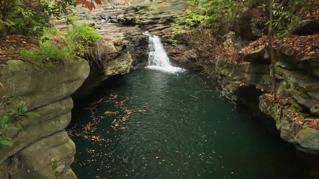 A small waterfall drops into a limestone ringed pool in a canyon while autumn leaves drift down in a gentle breeze.
