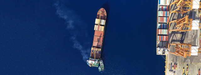 Aerial drone ultra wide top down photo of container ship cruising in Mediterranean port with deep blue sea assisted by tug boat