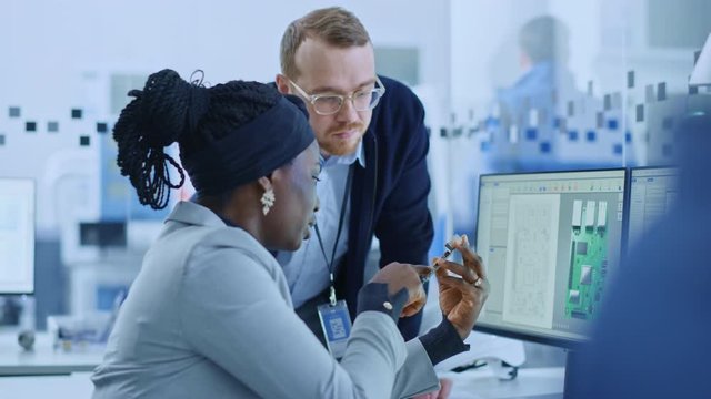 Modern Electronics Factory: Female Supervisor Talks to a Male Electrical Engineer who Works on Computer with CAD Software. Designing PCB, Microchips, Semiconductors and Telecommunications Equipment