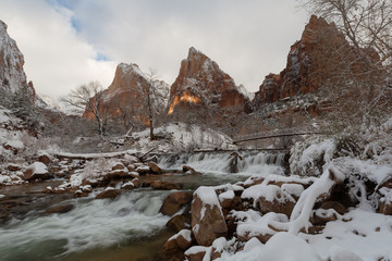 Obraz premium Snow covers the landscape in the Court of the Patriarchs in Zion National Park Utah while a small beam of sunlight breaks through the clouds to light a bit of the distant mountain.