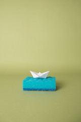 One blue sponge for washing dishes on a green background with an origami boat.