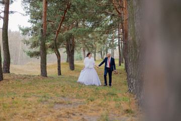 Happy bride and groom walk through the woods in autumn holding hands.