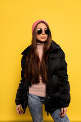 Fashion pretty carefree woman wearing a colorful pink cap and black jacket over yellow background. Appealing girl with long hair posing in studio