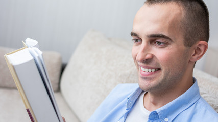 Fototapeta premium Guy reading a book sitting on sofa at home, close up, toned, 16:9