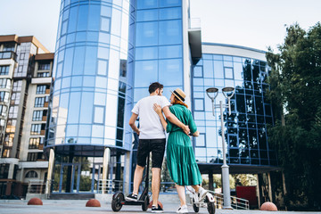 Man and woman on black and white electric scooters. Back view. Content technologies. Large modern building on background