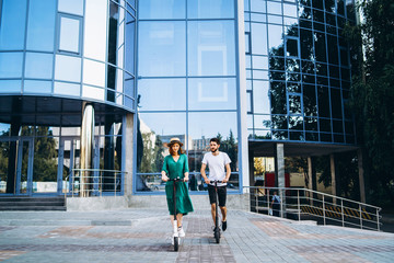 Full length portrait of a young romantic couple with electric scooters, walking in the city. Young woman in hat and man enjoy a walk