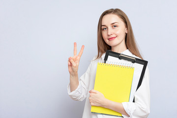 Naklejka premium Beautiful girl student with notebooks shows a peace sign. Concept of education. Portrait, grey background, copy space, slogan, toned