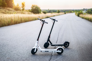 Two electric scooters, black and white, stand in the middle of the road at sunset time in the countryside. Content technologies. New movement