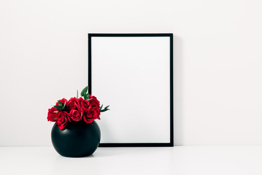 Red roses in black vase and an empty photo frame on background of white wall.