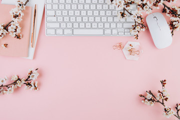 Spring flat lay top view home office workspace - modern keyboard and notebook with cherry blossom branches on a pink desk background