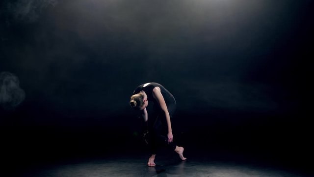 Beautiful elegant female in black leotard performing emotional modern contemporary dance in darkness of studio. Graceful young woman artist dancing contemp in twilight against black background