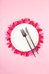 Round white plate with a black fork and a knife on a pink table with paper hearts. Valentine's day concept and laid table.