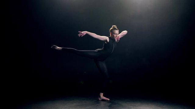 Pretty young actress performing expressive dance elements against black background. Beautiful fit female in black lights dancing modern contemporary choreography in dusk of dance studio
