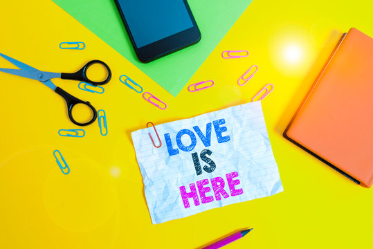 Text sign showing Love Is Here. Business photo text Roanalysistic feeling Lovely emotion Positive Expression Care Joy Paper sheets pencil clips smartphone scissors notebook colored background