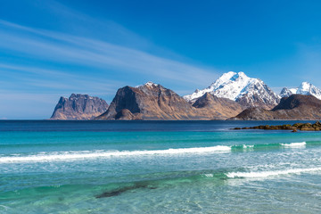 Fototapeta na wymiar Beautiful beach during springtime on a sunny day with blue sky and silent ocean with small waves. Mountain chain with snow in the background. From Myrland beach in Lofoten - Norway. Blue clear water.