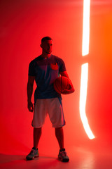 Young athletic man, basketball player stands holding ball in one hand with back lights and red background