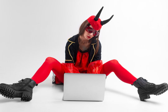 Sexy brunette woman in red tights, black boots and devil horns using laptop over white studio background.