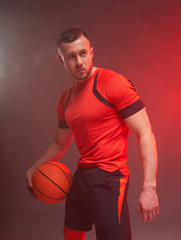 Young athletic man, basketball player posing holding ball in one hand with back lights and smoke on...