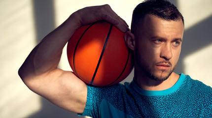 Portrait of basketball player holding ball on shoulder and sitting near wall with shadows from window